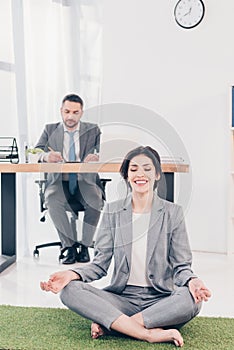 Smiling businesswoman meditating in Lotus Pose on grass mat while businessman sitting at table and writing at desk in