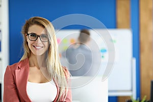 Smiling businesswoman posing in office with in presentable pink costume, colleague on background