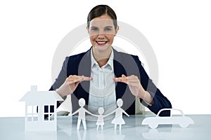 Smiling businesswoman protecting paper cut out family, house and car with hands
