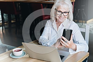 Smiling businesswoman is sitting at table in front of laptop and using smartphone. Pensioner freelancer works.