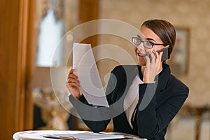 Smiling businesswoman standing in expensive hotel room, looking at documents and talking by phone