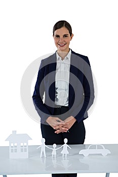 Smiling businesswoman standing with paper cut out of family, house and car on table