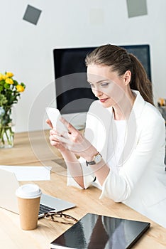 smiling businesswoman in stylish suit using smartphone at workplace