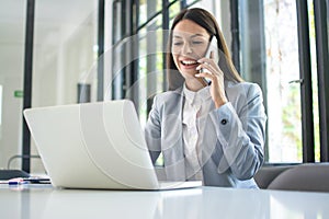 Smiling businesswoman talking on mobile phone and using laptop in office.