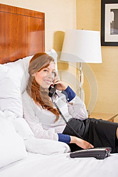 Smiling businesswoman talking on phone while sitting on the bed in hotel room.