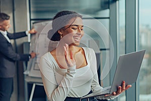 Smiling businesswoman talking via video call with client and waving hi while standing in office