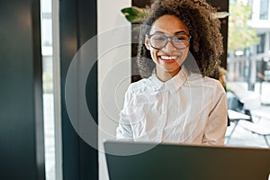 African businesswoman working laptop while standing near window in coworking and looking camera