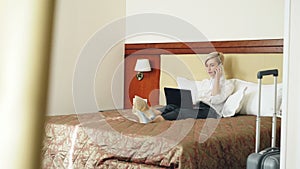 Smiling businesswoman in white shirt using on laptop and talking at mobile phone while lying on bed in hotel room