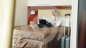 Smiling businesswoman in white shirt using on laptop and talking at mobile phone while lying on bed in hotel room