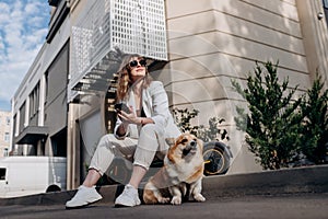 Smiling businesswoman in white suit sitting on electric scooter and using phone during walking with Welsh Corgi Pembroke dog in