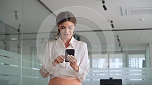 Smiling busy business woman of middle age using mobile phone standing in office.