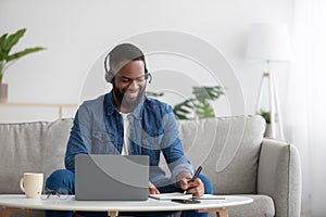 Smiling busy young black businessman freelancer or student with beard in headphones with pc making notes, study