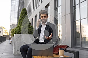 Smiling busy young european man manager with beard in suit with cup of coffee and laptop calls by phone