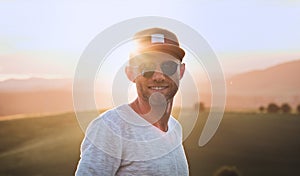Smiling at camera handsome cheerful man with 3-Day Stubble Beard, baseball cap and fancy blue sunglasses with bright sunset sun photo