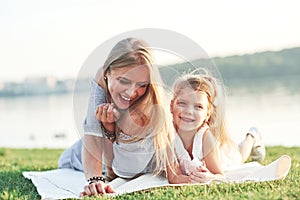 Smiling at the camera. Photo of young mother and her daughter having good time on the green grass with lake at