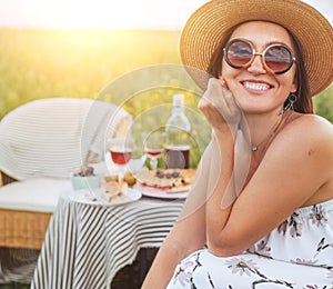 Smiling at camera young Sincerely  Woman in sunglasses and straw hat dressed light summer dress sitting in rattan chair on the