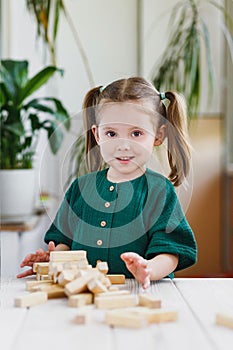 Smiling canny cute child in green dress and broken wooden jenga tower on a table. Indoor activities concept