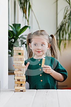 Smiling canny cute child in green dress looking at a wooden jenga tower standing on a table