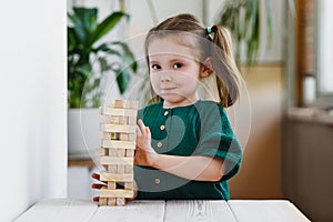 Smiling canny cute child in green dress touching a wooden jenga tower standing on a table