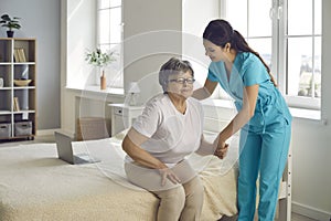 Smiling caring nurse supporting patient assisting senior woman get up from bed