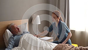 Smiling caring young female doctor helping senior adult male patient to lying on bed and covering him blanket at home