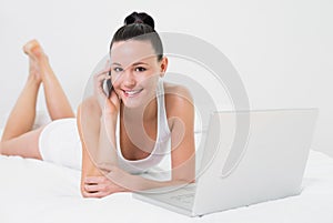 Smiling casual woman using cellphone and laptop in bed