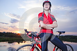 Smiling Caucasian Bicyclist Made on Sunset Golden Hour Time. Posing with Road Bike and Equipped with Professional Outfit