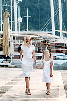 Smiling caucasian blonde  mother and daugher walking by harbor with yachts on a touristic sea resort