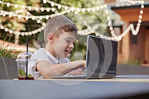 smiling caucasian boy sitting at table at veranda of cafe, studying with laptop