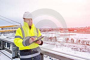A smiling caucasian builder in a white hard hat and yellow fluorescent jacket holds clipboard