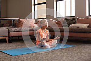 Smiling caucasian elementary girl mediating while sitting on yoga mat by sofa