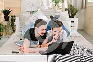 Smiling Caucasian gay couple using laptop on bed at home. Homosexual, gay couple concept