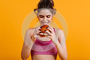 Smiling caucasian girl eats burger. Indoor portrait of amazing woman with sandwich
