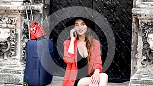 Smiling caucasian girl with a suitcase is talking on cell phone outdoors.