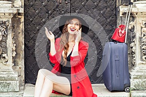 Smiling caucasian girl with a suitcase is talking on cell phone outdoors.