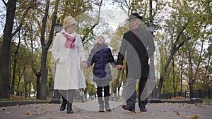 Smiling Caucasian girl walking with her grandparents along the alley in the park. Senior couple dressed in elegant