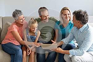 Smiling caucasian grandparents on couch with granddaughter and her parents looking at tablet
