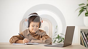 Smiling caucasian little kid in headphones sits at table, makes notes, watches lesson on computer in room