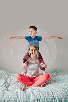Smiling Caucasian mother and boy son playing in bedroom at home. Child sitting on moms shoulders and laughing. Family having fun