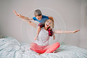 Smiling Caucasian mother and boy son playing in bedroom at home. Child sitting on moms shoulders and laughing. Family having fun