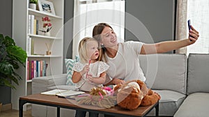 Smiling caucasian mother and little daughter, sitting on sofa at home, making memories together, drawing on notebook and taking