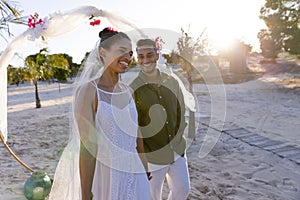 Smiling caucasian newlywed couple standing at beach at wedding ceremony against sky at sunset