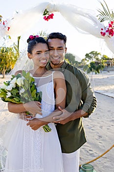 Smiling caucasian newlywed young couple with bouquet standing at beach at wedding ceremony
