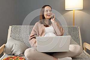 Smiling Caucasian pregnant woman using laptop and talking on cellphone resting on sofa having phone conversation with friend while