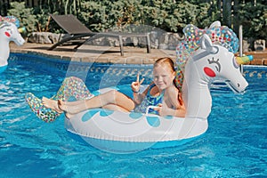 Smiling Caucasian red-haired  girl lying on inflatable ring unicorn. Kid child enjoying having fun in swimming pool. Summer