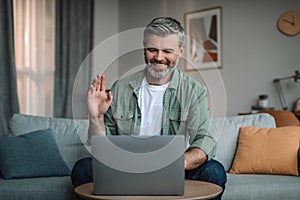 Smiling caucasian retired man with beard waves hand, looks at laptop in living room interior