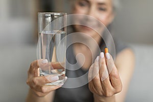 Smiling caucasian retired woman show glass of water and pill of vitamins, painkillers or antidepressants in room