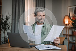 Smiling caucasian senior man with beard works on laptop with documents in living room interior