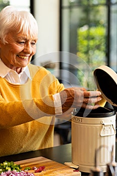 Smiling caucasian senior woman preparing food, composting vegetable waste in kitchen