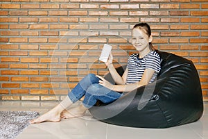 Smiling caucasian teen girl sitting in black bean bag chair against brick wall. Casual outfit. Childhood concept. Blank white scre